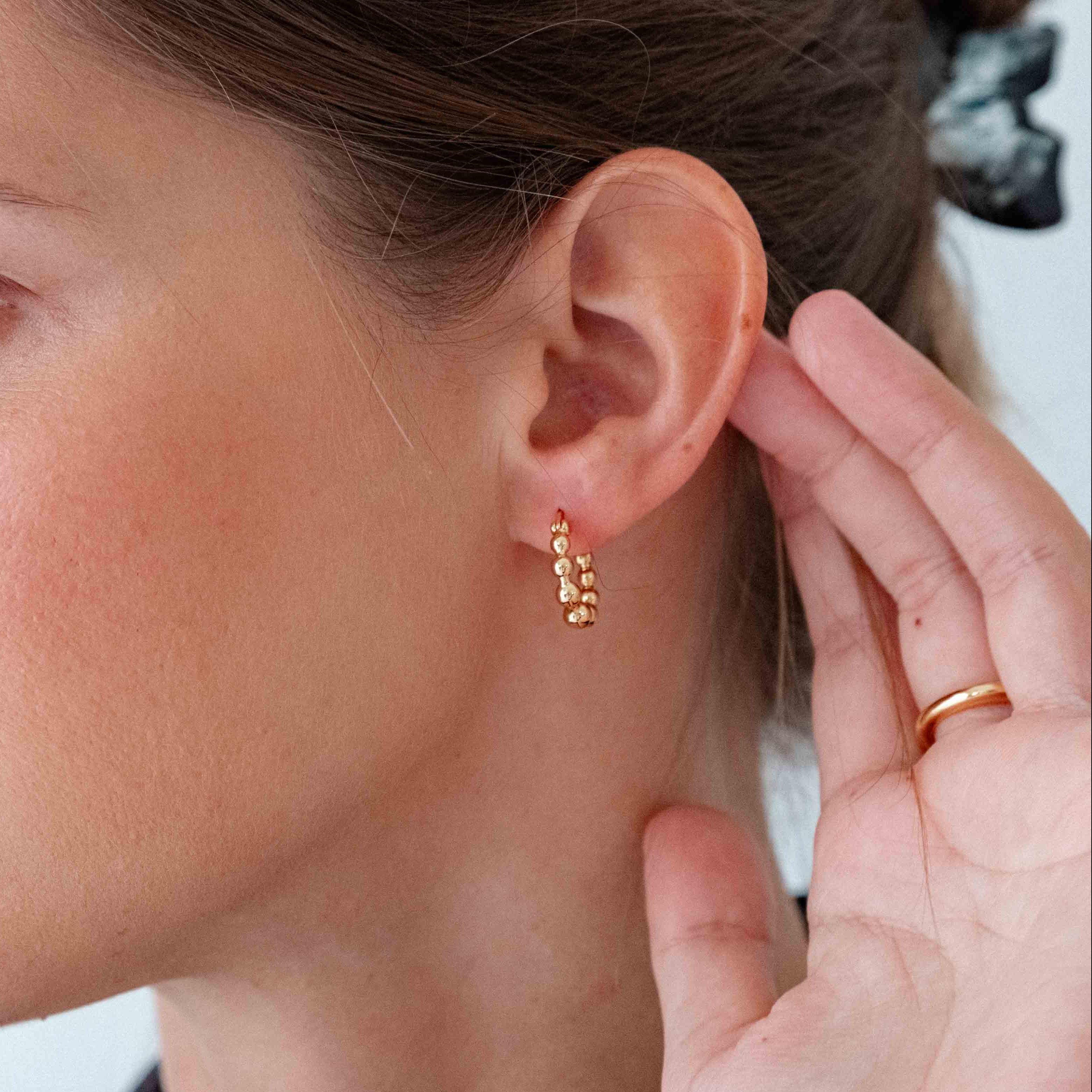 Woman wearing gold earrings with a hand adjusting them, against a neutral background