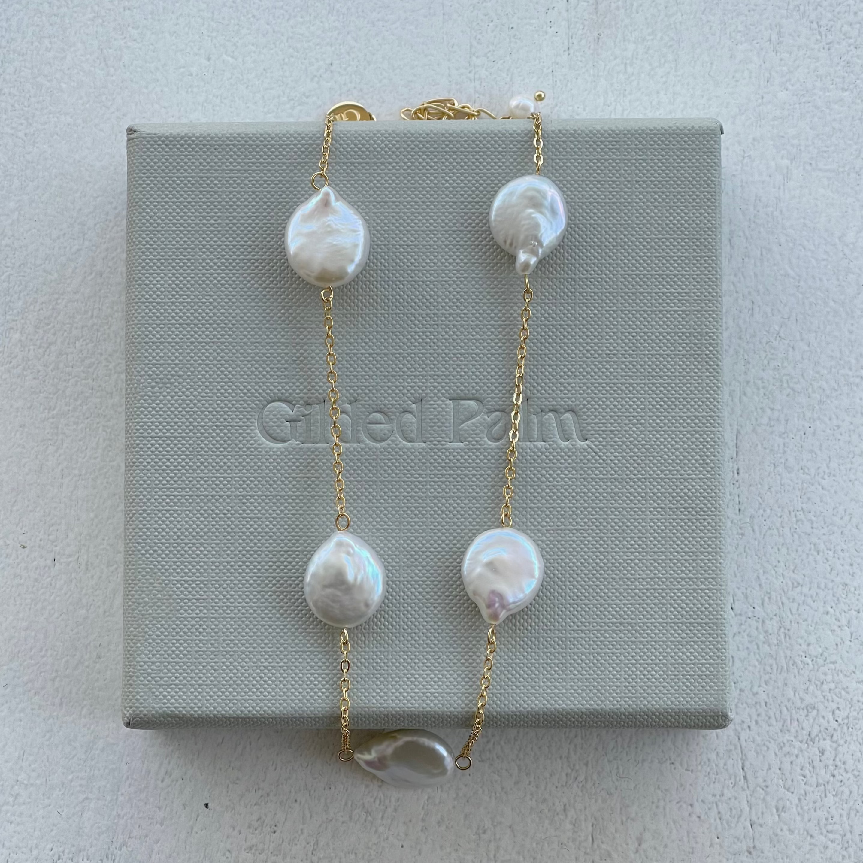 A gold necklace with baroque pearls displayed on a grey box with the brand 'Gifted Palm' inscribed.