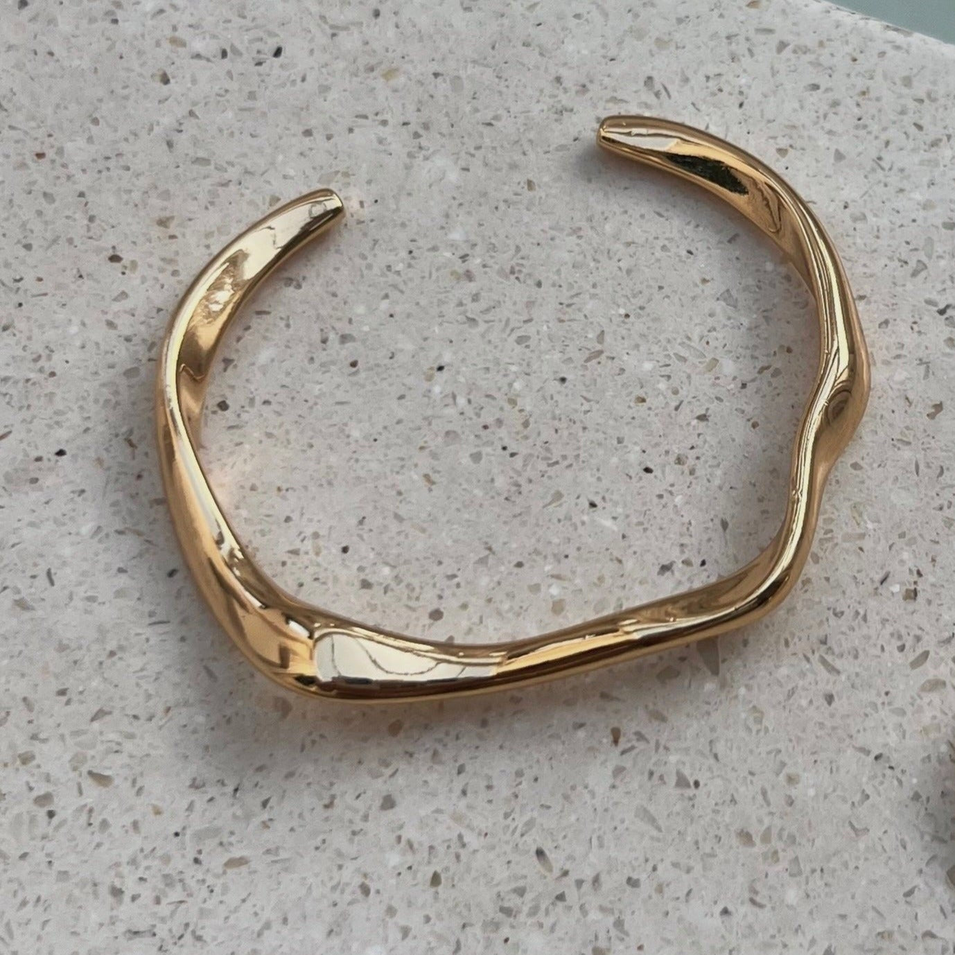 A gold hook bracelet displayed on a textured surface, with a close-up inset showing the bracelet's gold plated and design.